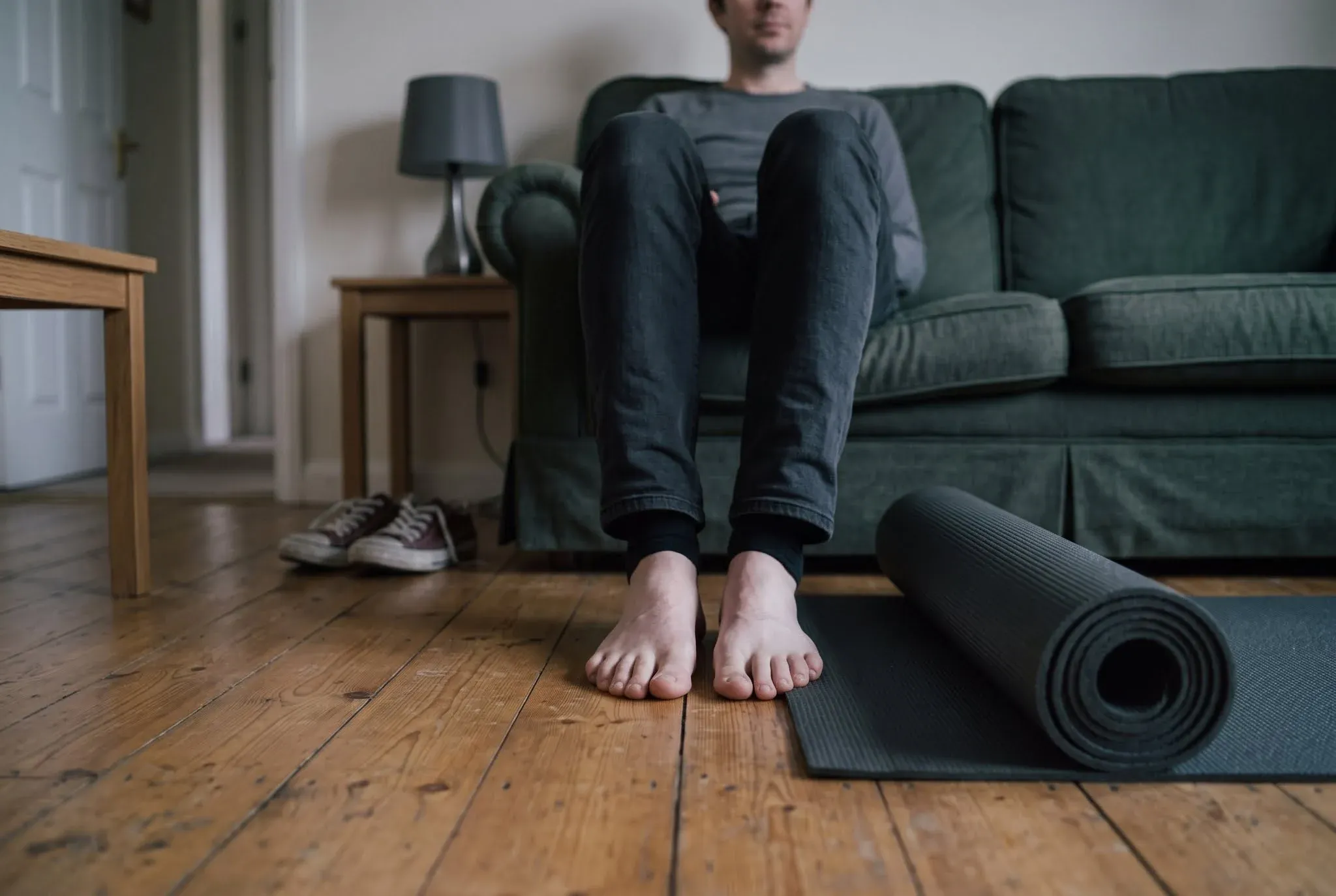 Bare feet resting on a wooden living room floor beside a rolled exercise mat, a man seated on the sofa behind them