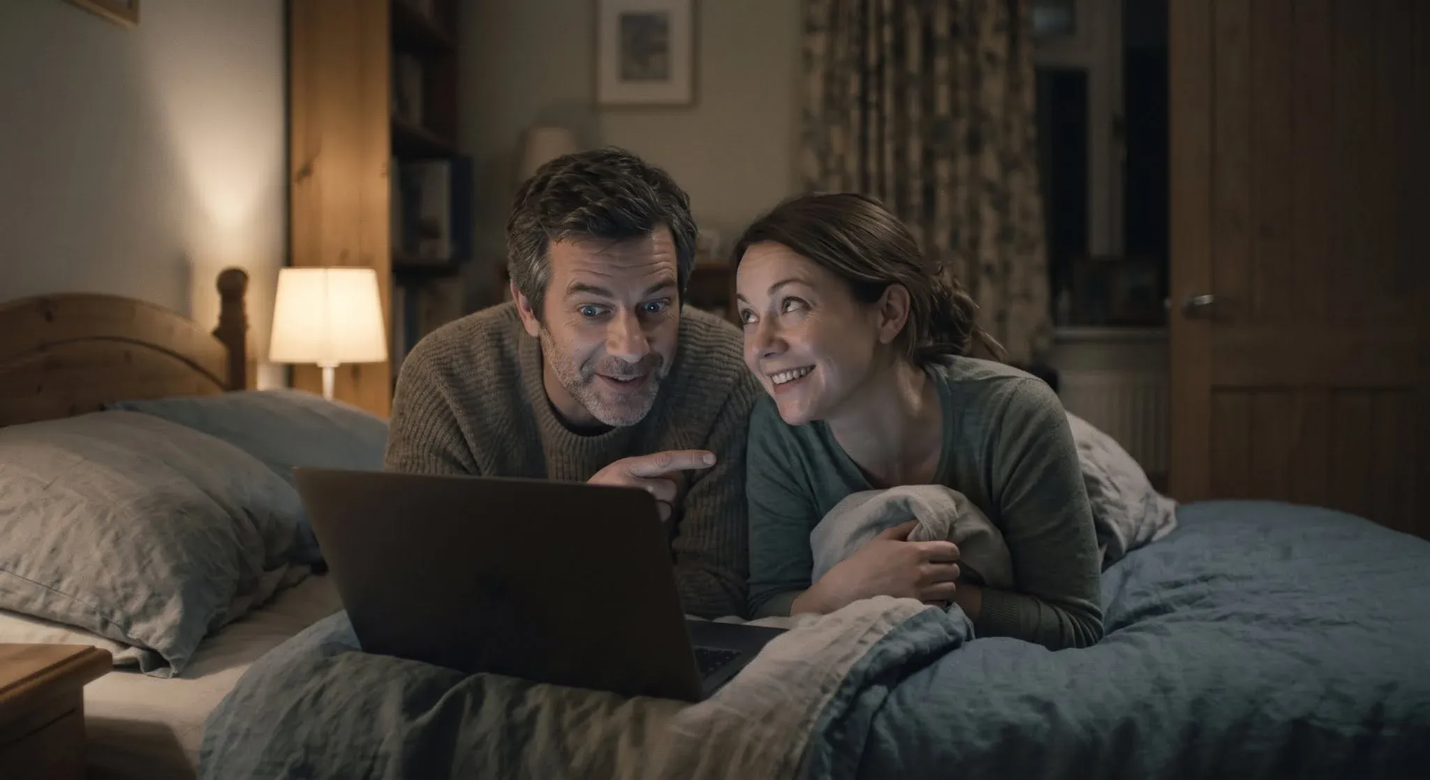 A couple lying on a bed looking at a laptop together, the man pointing excitedly at the screen and the woman smiling, hopeful