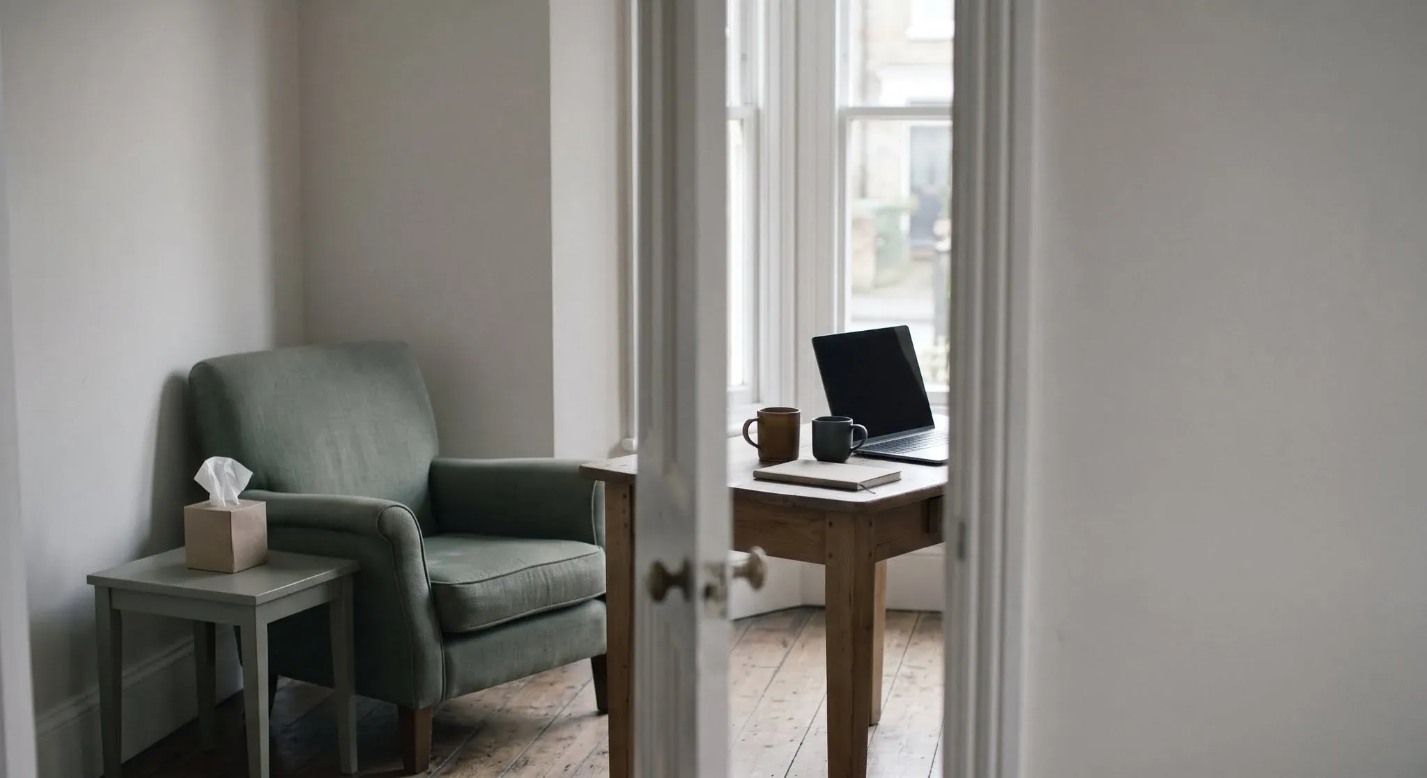 A quiet room with a green armchair and a box of tissues on one side of a doorway, and a desk with a laptop and two mugs visible through it