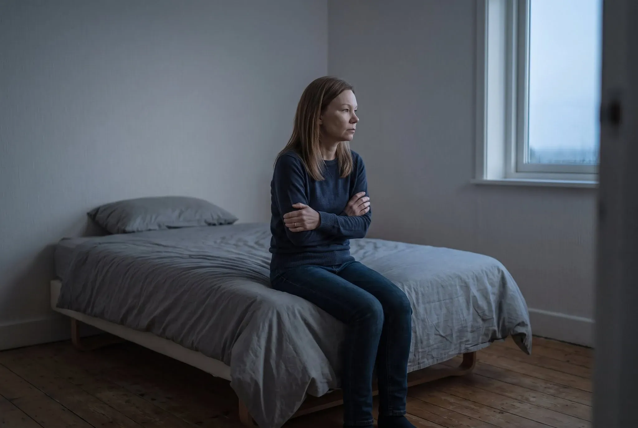 A woman sitting fully dressed on the edge of a made bed with her arms folded, looking toward a window in cold morning light