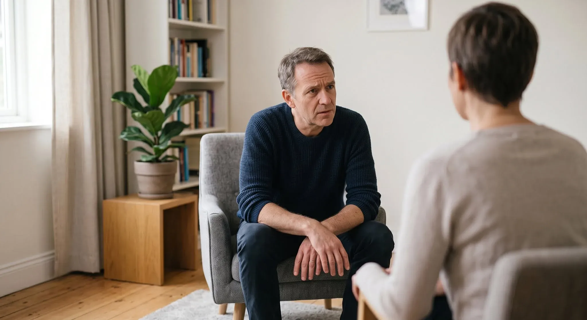 A man in his fifties leaning forward in an armchair during a therapy session, listening with a troubled expression, the therapist seen from behind