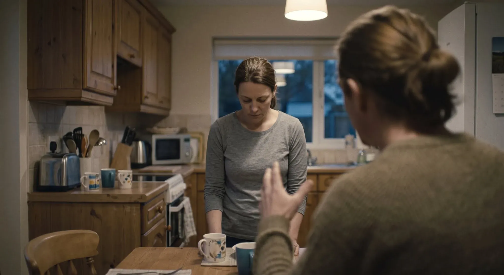 A woman standing very still at a kitchen counter, looking down, while a partner in the foreground gestures sharply mid-sentence