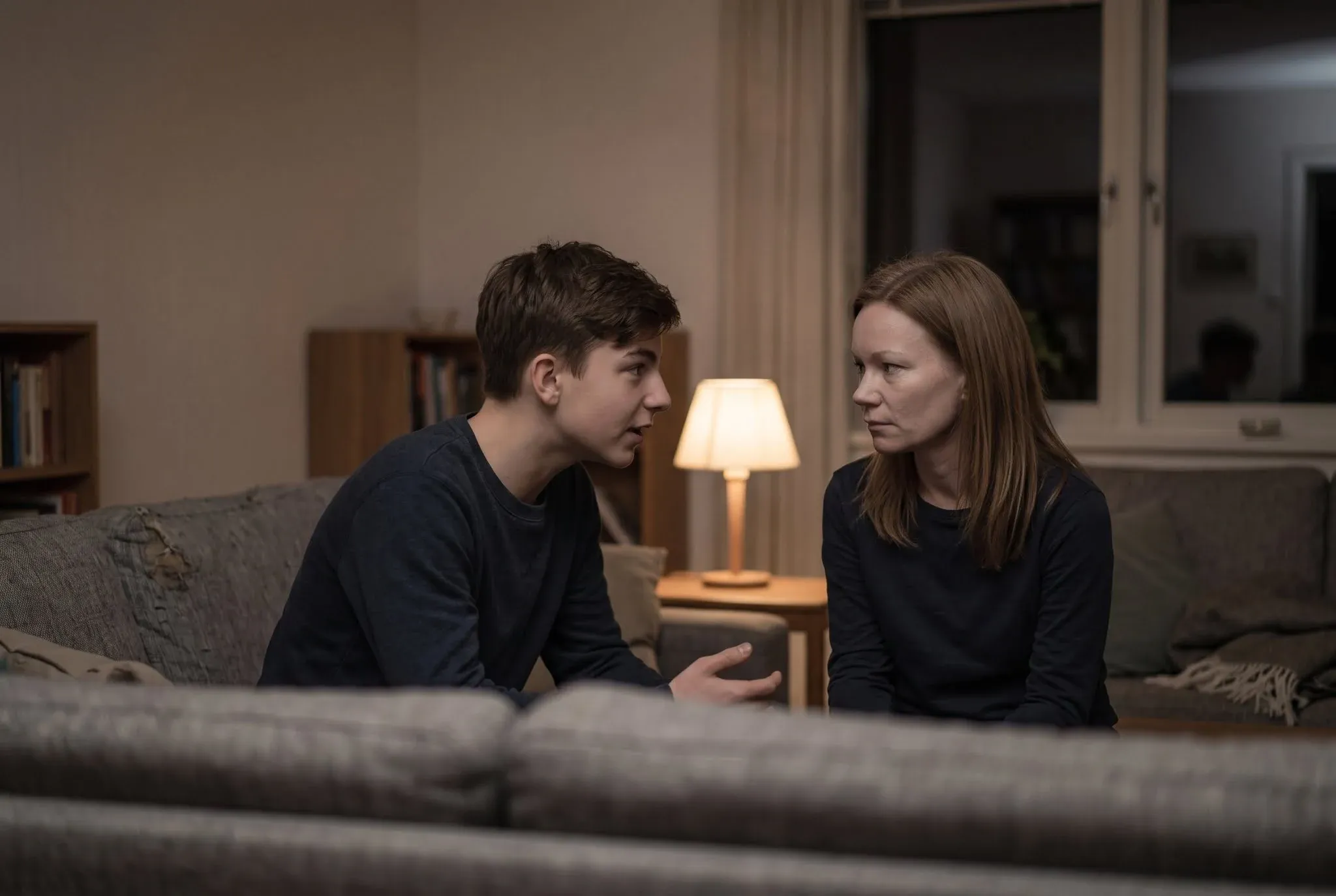 Two people sitting close on a sofa in lamplight, one talking and gesturing while the other listens with a guarded expression