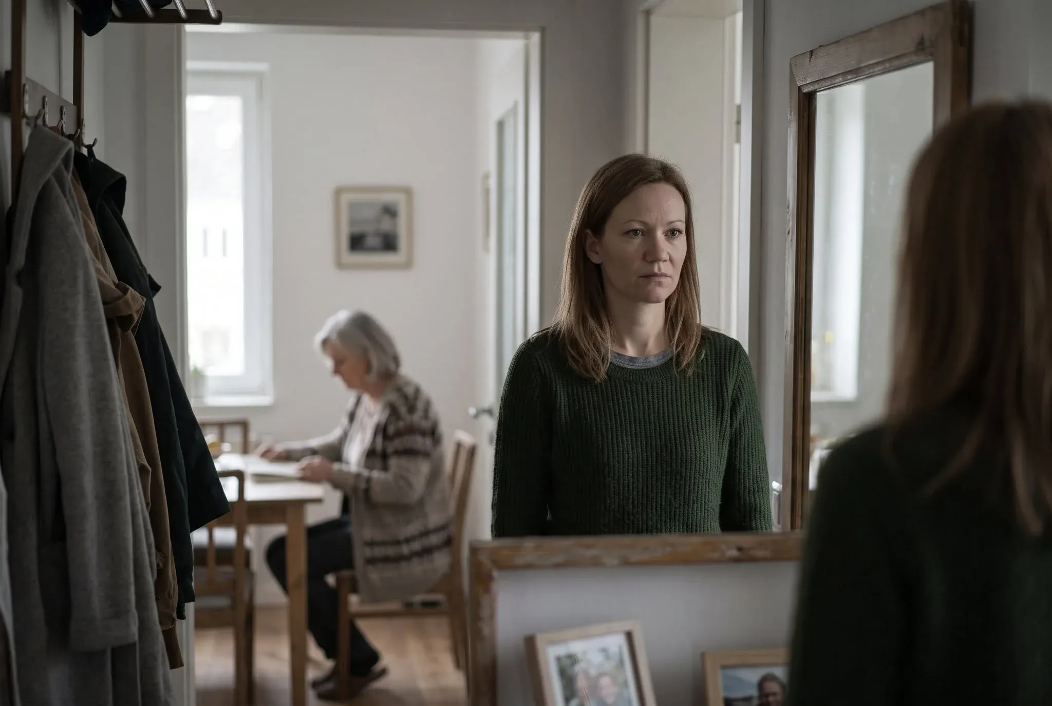 A woman looking at her reflection in a hallway mirror while an older woman sits at a table in the room behind her