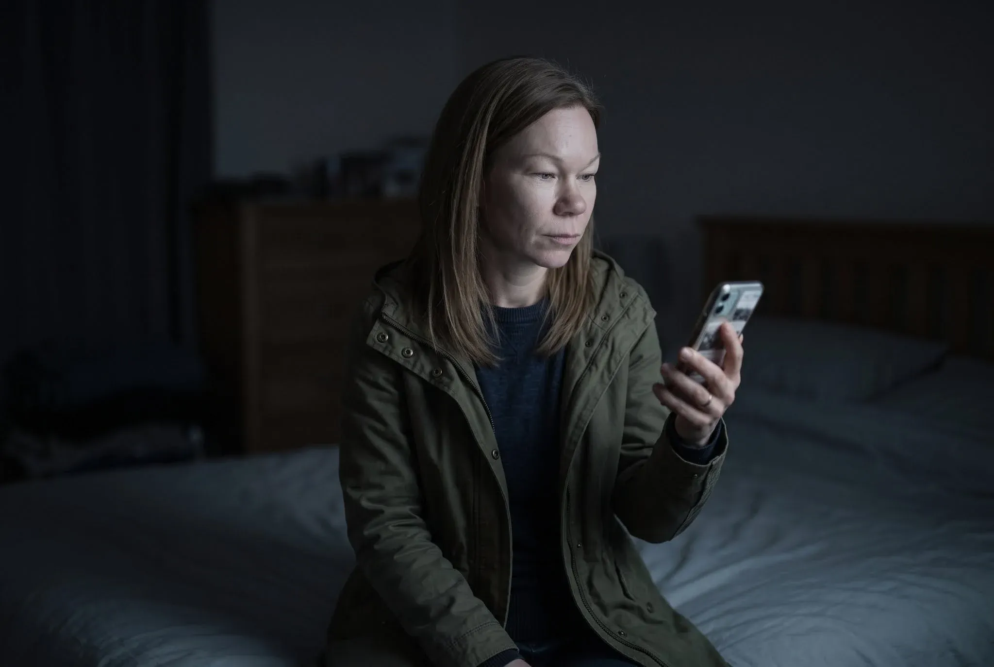 A woman sitting on the end of a bed in a dark room, still in her coat, reading her phone by its light