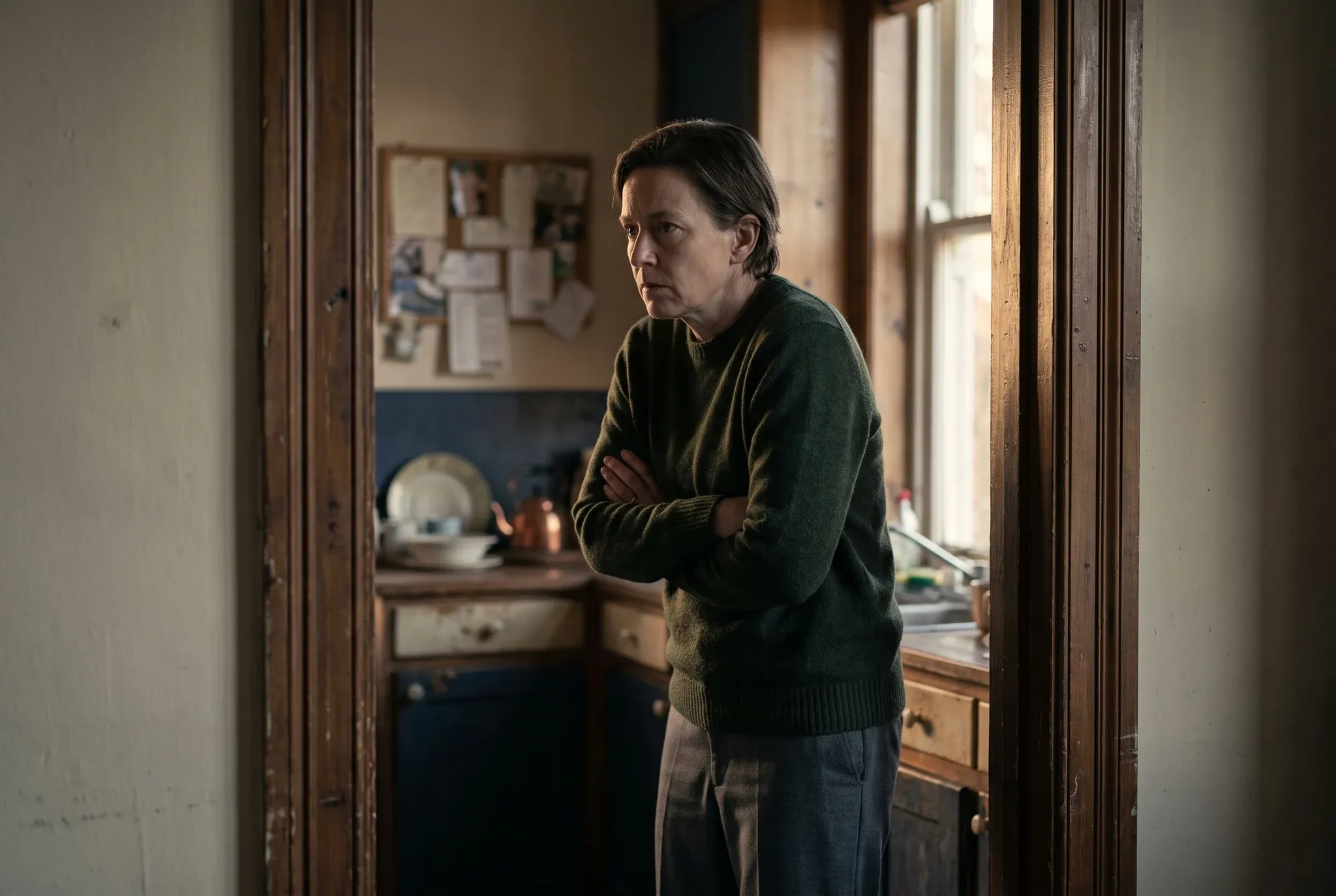 A woman standing tensely in a kitchen doorway with her arms folded, looking away