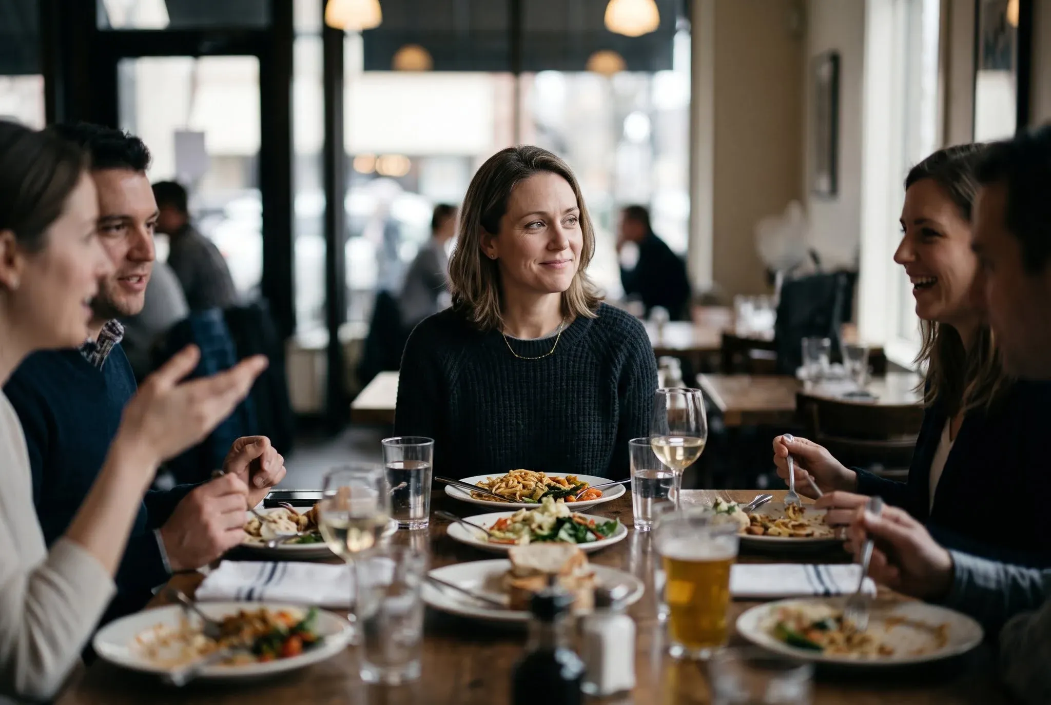 A woman at a busy restaurant table smiling politely and looking past the conversation, her plate untouched