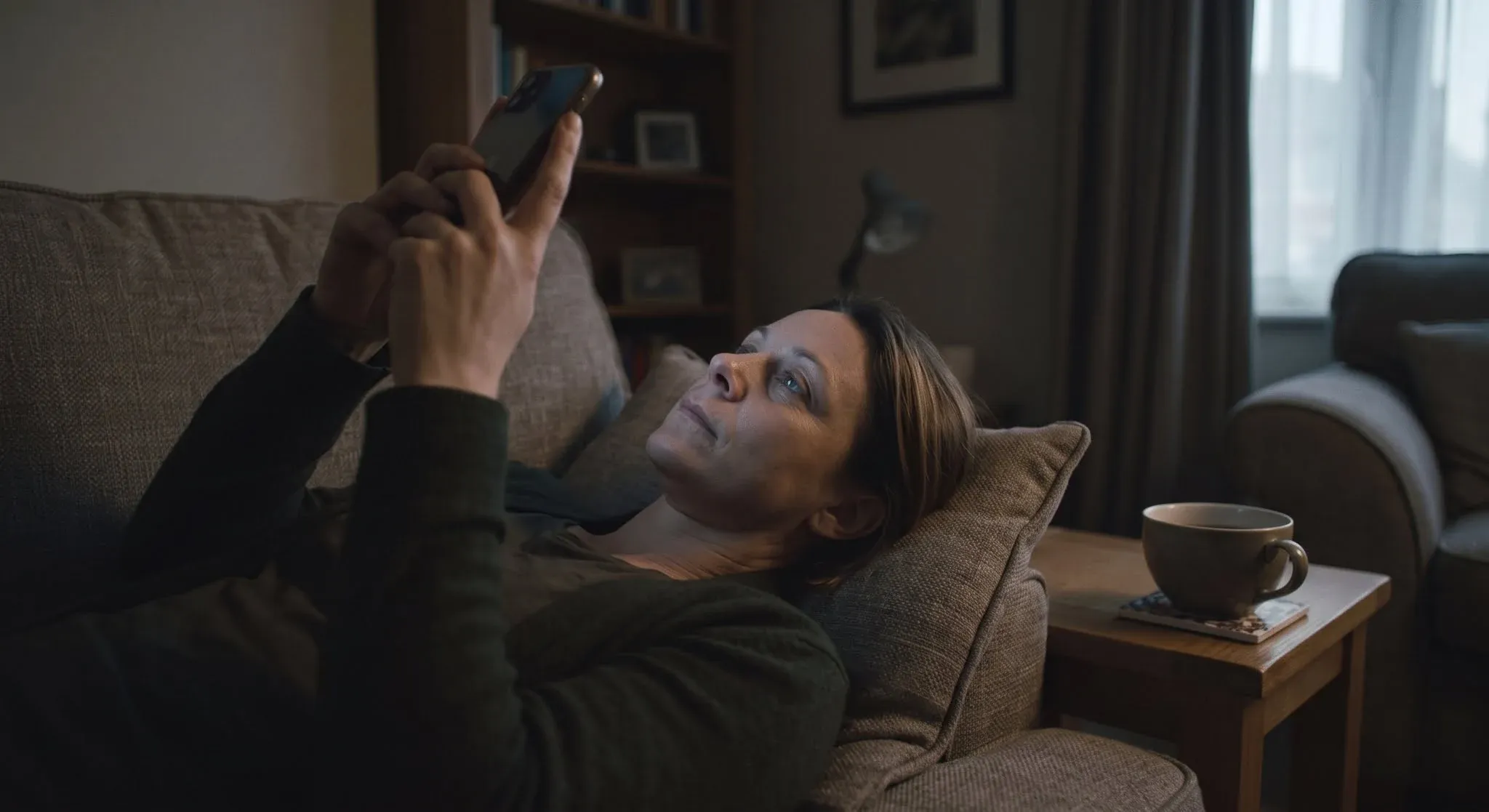 A woman lying on a sofa in a dim living room, holding her phone above her face and waiting, a cold cup of tea on the side table