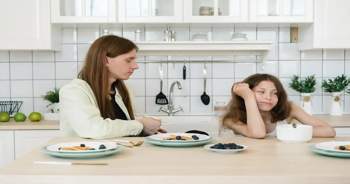 A woman and a teenage girl sitting at a kitchen table with untouched plates between them, neither looking at the other