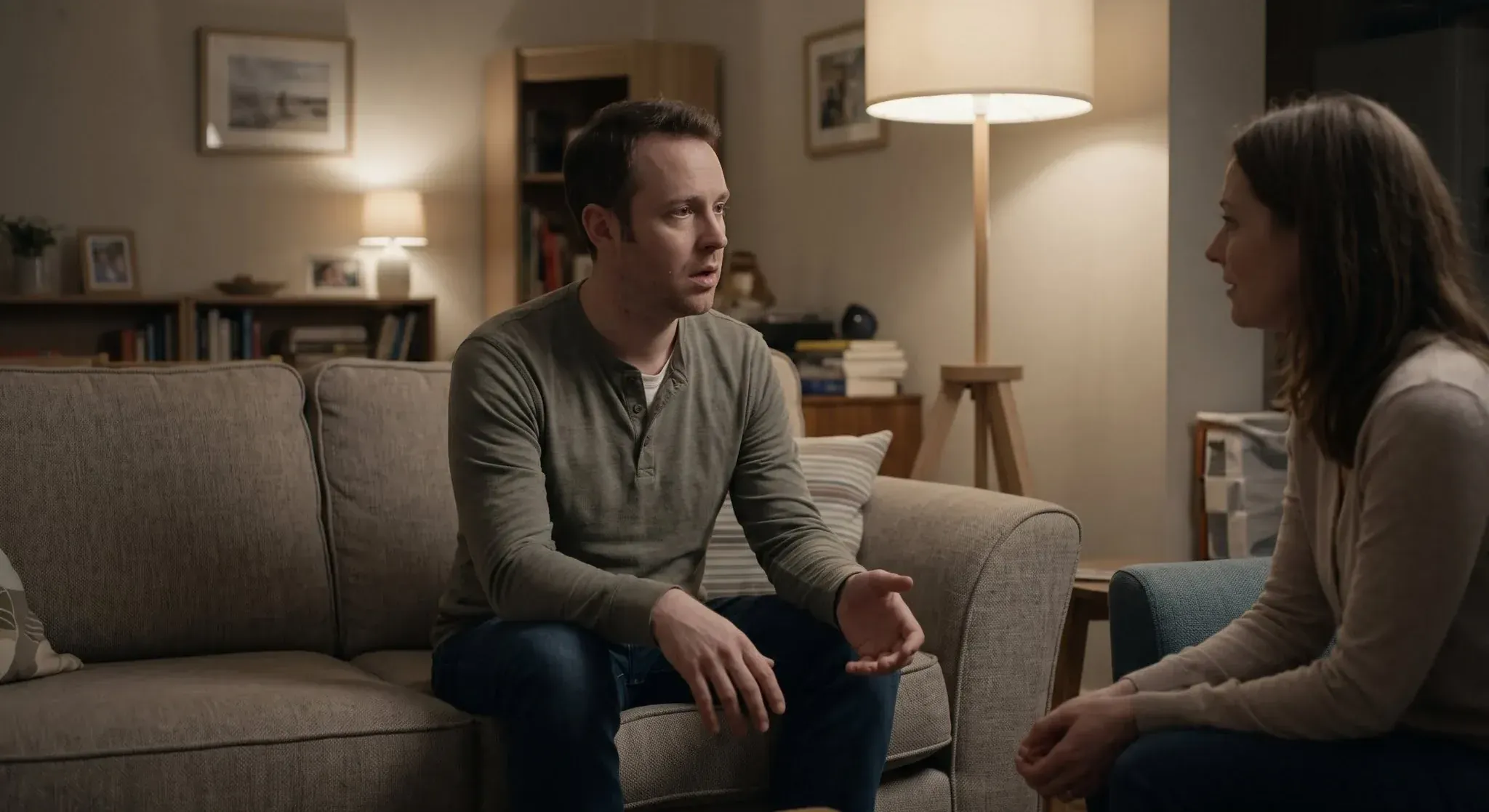 A man on a sofa in a lamp-lit living room, searching for words to answer a woman who is listening to him
