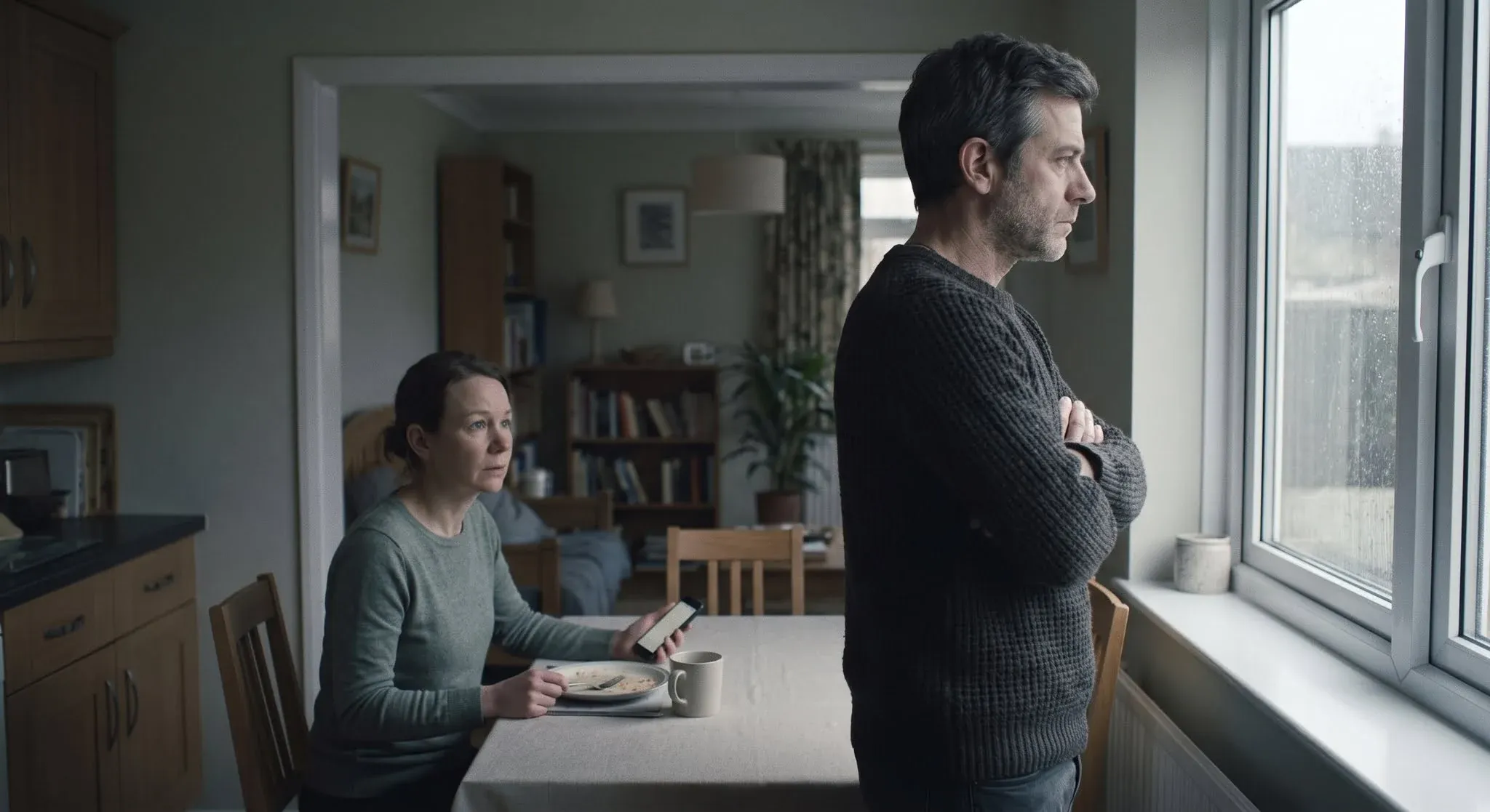 A man standing rigid at a kitchen window with his arms folded and his back to the room, while a woman at the table behind him looks towards him uncertainly, phone in hand