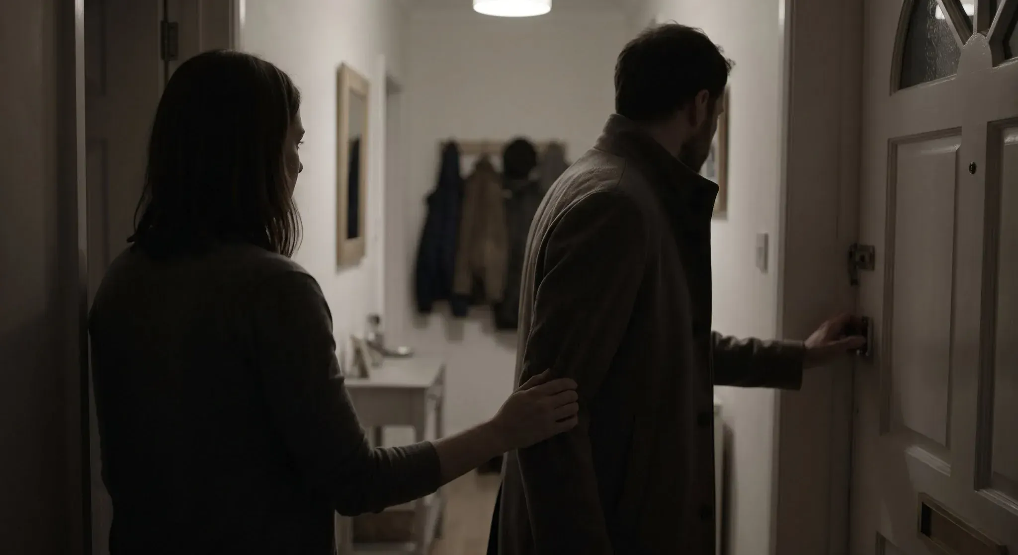 A woman in a dim hallway at night holding the sleeve of a man in a coat whose hand is already on the front door latch