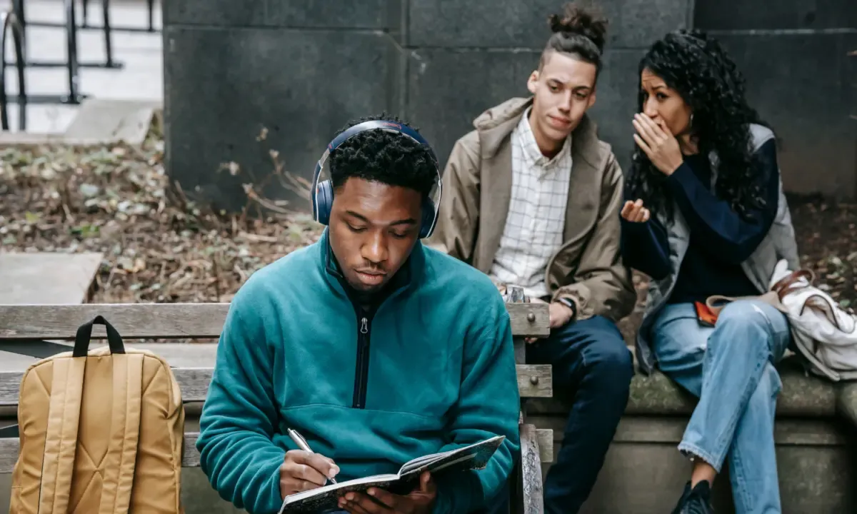 A young man writing in a notebook while two people behind him talk with a hand raised to the mouth