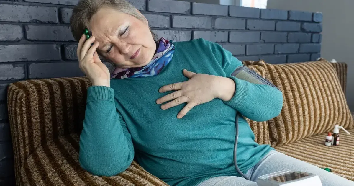 An older woman sitting on a sofa with one hand pressed to her chest, a phone beside her