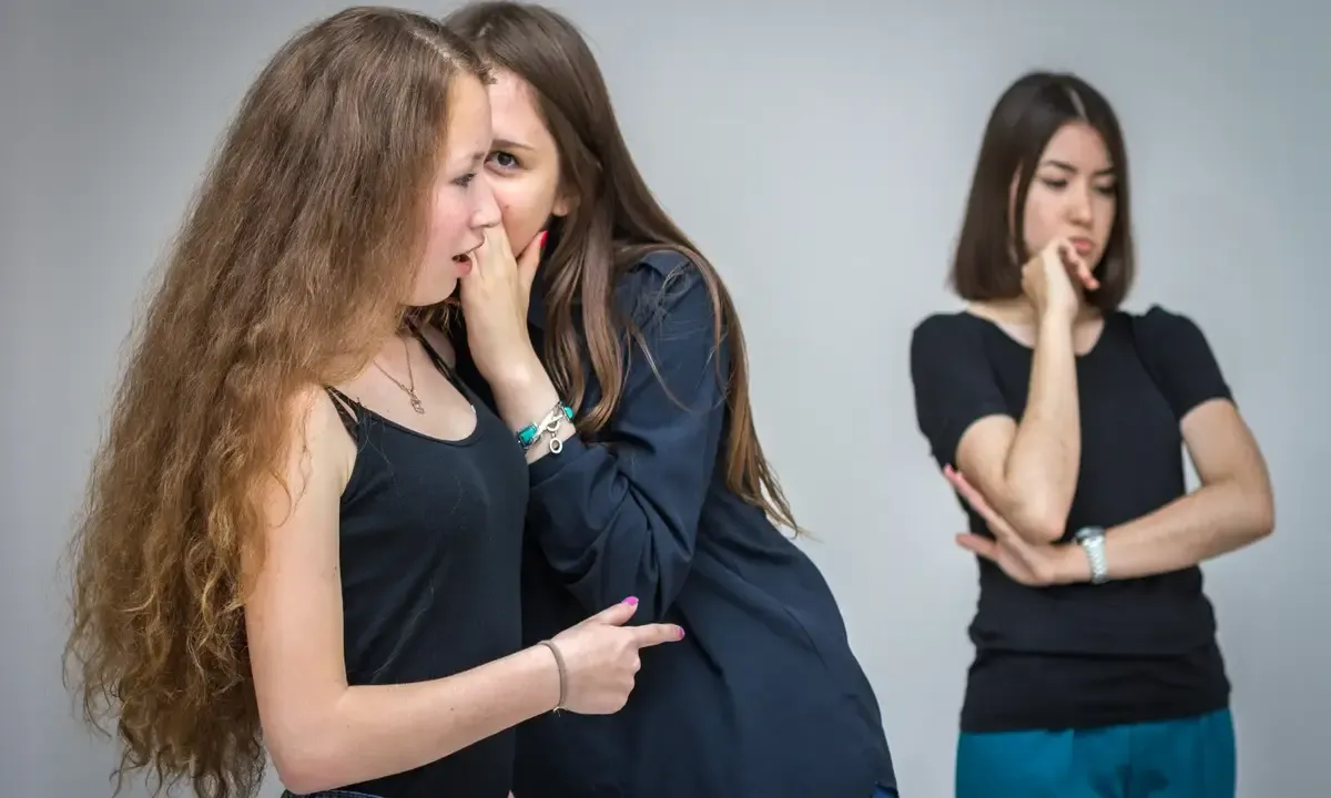 Two women whispering together while a third woman stands apart with her arms folded