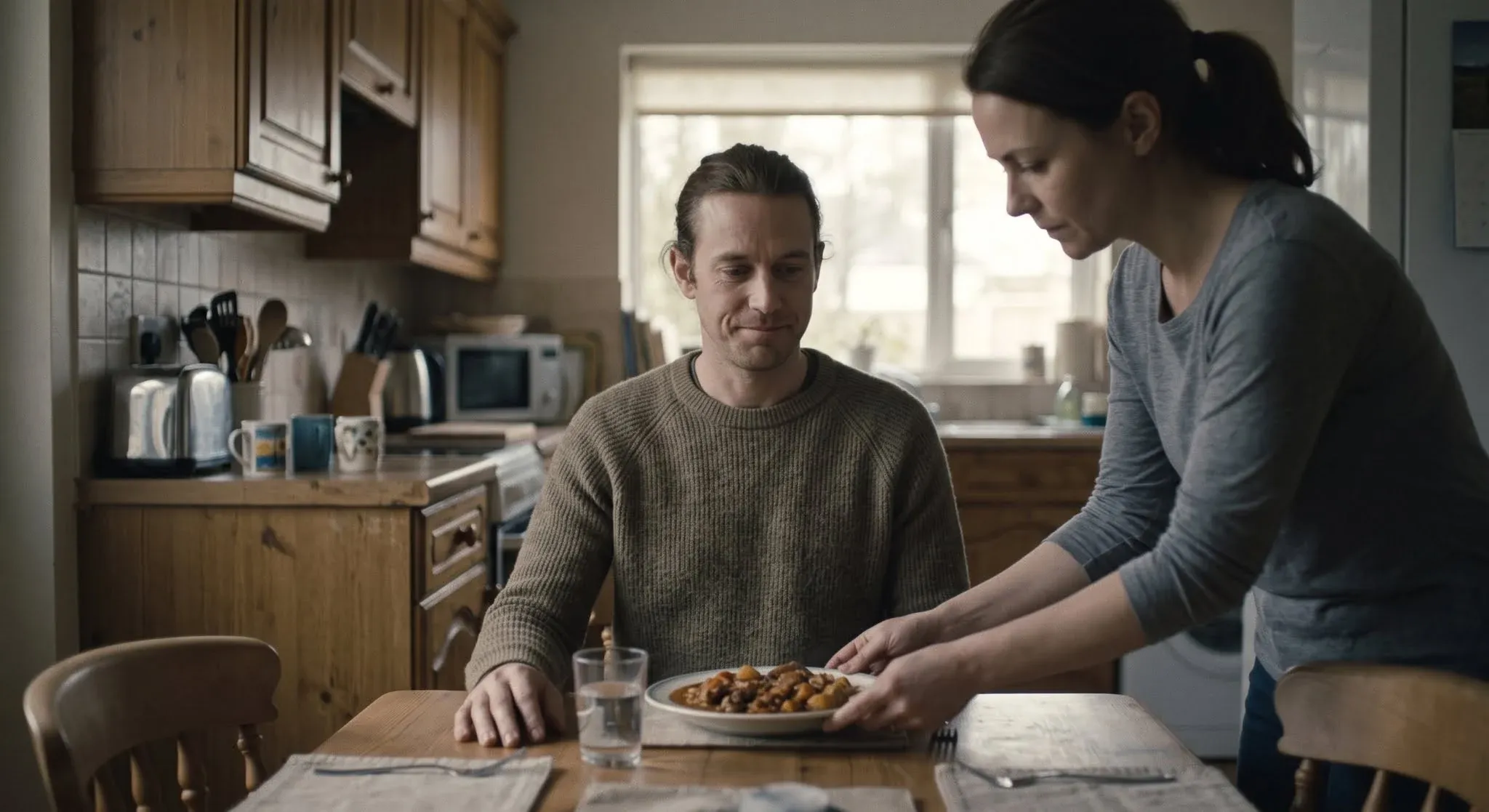 A man at a kitchen table with a slightly awkward smile as a woman sets a plate of food down in front of him
