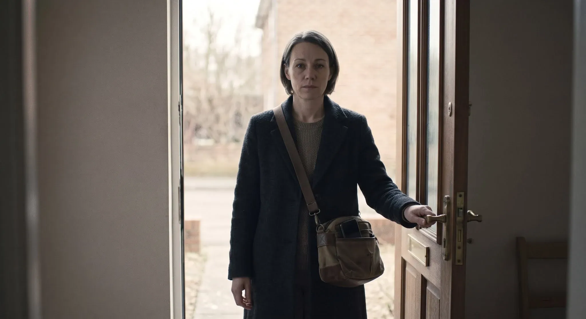 A woman in a coat with a bag over her shoulder standing calmly at an open front door, hand on the handle, about to leave