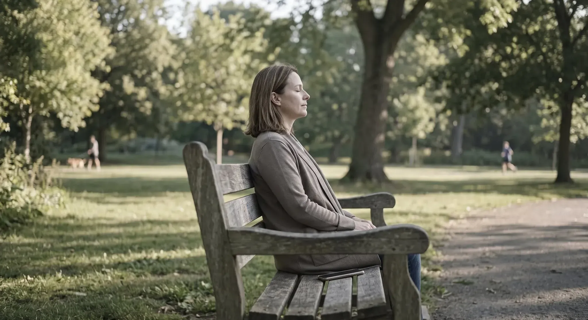 A woman sitting on a park bench in the morning with her eyes closed, her phone lying face down on the bench beside her