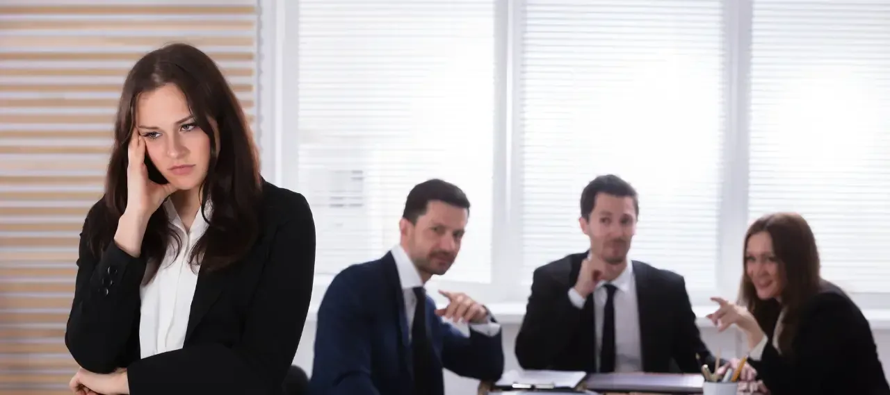 A woman looking distressed in the foreground while colleagues talk behind her at a table